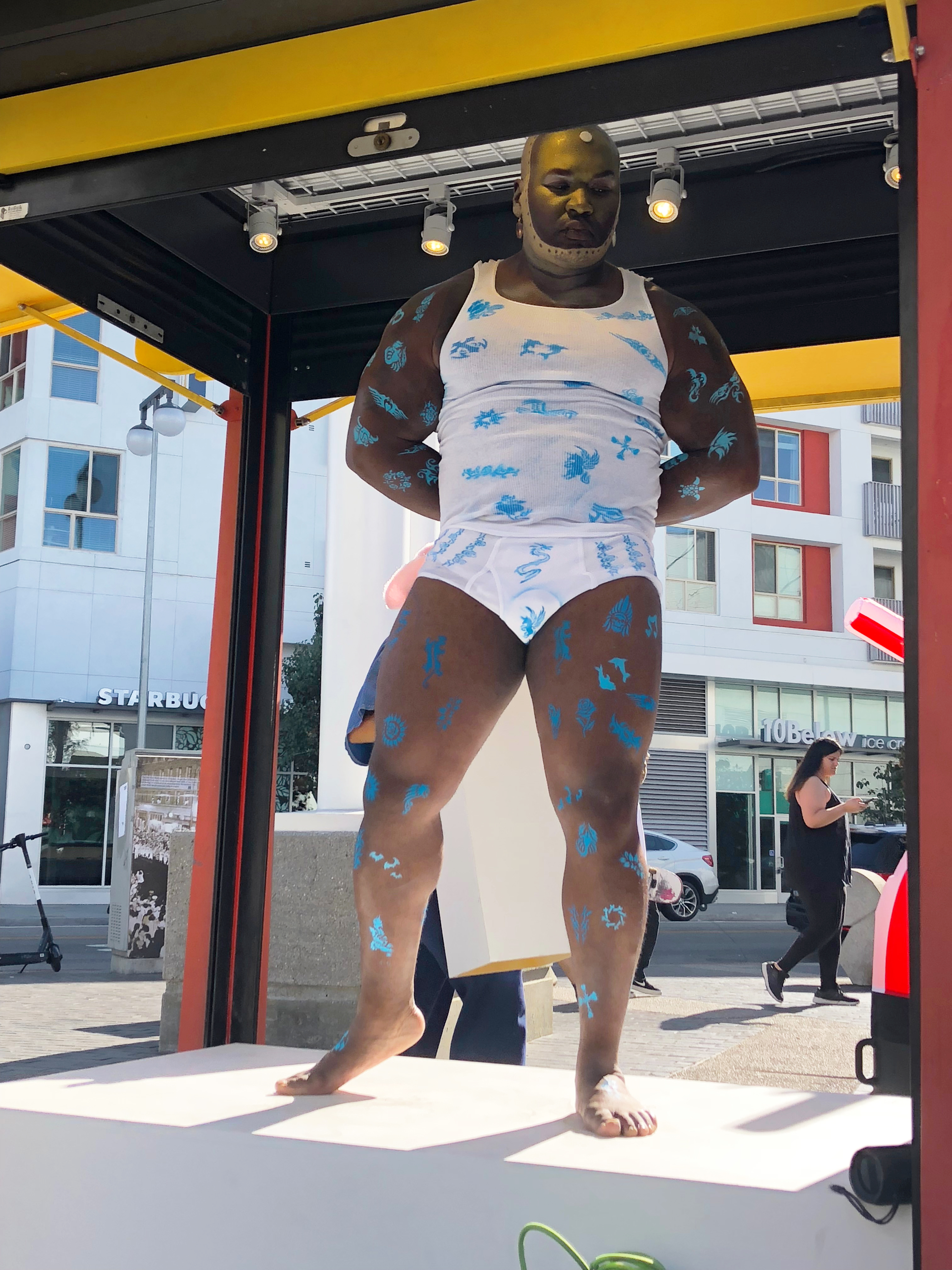 A muscular black man in tank top and underwear posing outside in a cell phone kiosk 